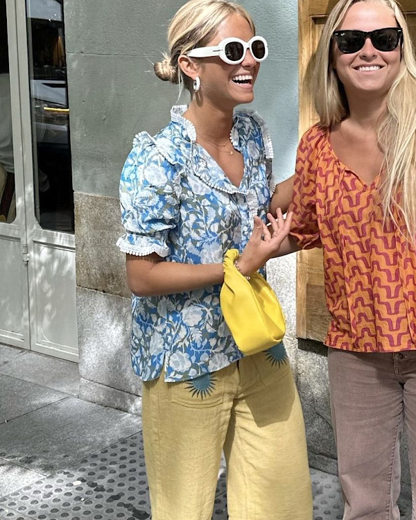 Femme portant une blouse fleurie bleu clair, un pantalon jaune pâle et des lunettes blanches en ville