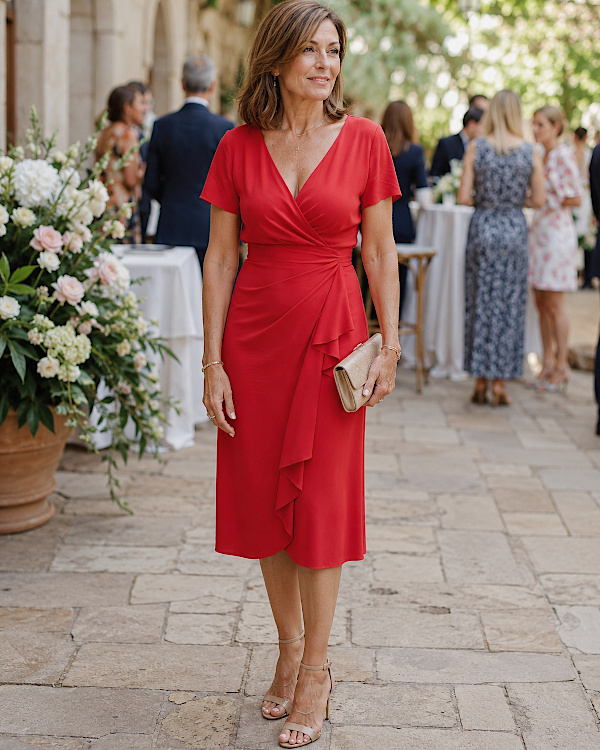 Femme élégante portant une robe rouge portefeuille avec pochette beige et sandales dorées lors d’un mariage