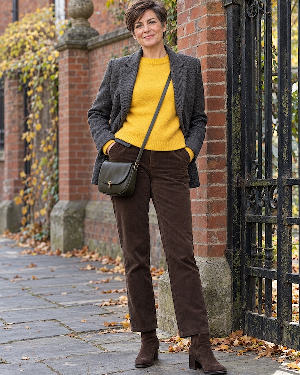Femme aux cheveux courts portant un pull jaune, un blazer gris, un pantalon en velours marron, des bottines et un sac en bandoulière